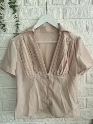 Beige blus med djup v-ringning - Stilren beige blus med korta ärmar och djup v-ringning. Blusen har knappar framtill och fina veckdetaljer över bysten som ger en snygg siluett. Tillverkad i ett mjukt, tunt tyg som känns luftigt och bekvämt. Perfekt till jeans eller kjol.