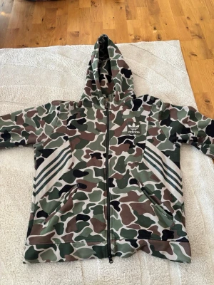 Adidas camo hoodie med dragkedja - Säljer en Adidas hoodie med grön, brun och svart camomönster. Tröjan har dragkedja framtill, två fickor och klassiska vita Adidas-ränder på sidorna. Stor huva och Adidas-logga på bröstet. Perfekt för dig som gillar streetwear och vill sticka ut.