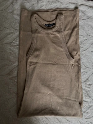 Beige ribbad långklänning från ZARA - Säljer en beige ribbad långklänning från ZARA med ärmlös design och rund halsringning. Klänningen har en slim passform och är tillverkad i mjuk bomullsmix, perfekt för en stilren och enkel look.