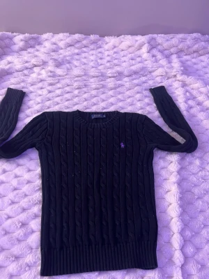 Svart kabelstickad tröja Polo Ralph Lauren - Kabelstickad svart tröja från Polo Ralph Lauren med lila broderad logga på bröstet. Tröjan har rund halsringning, långa ärmar och ribbade muddar vid ärmslut och nederkant. Perfekt för en stilren och klassisk look.