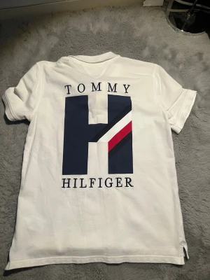 Vit piké från Tommy Hilfiger - Säljer en vit pikétröja från Tommy Hilfiger med stor blå H-logga och röd detalj på ryggen. Klassisk krage och knappar framtill. Perfekt för dig som gillar stilrena och sportiga plagg med tydlig logga. 8/10 skick