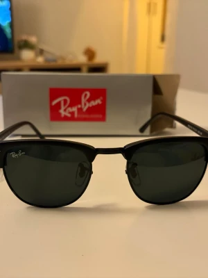 Svarta Ray-Ban solglasögon - Ray-Ban solglasögon med klassisk svart båge och mörka linser. Modellen har en stilren och tidlös design med metallbåge och Ray-Ban-logga på linsen. Perfekt för dig som vill ha en ikonisk look och skydda ögonen mot solen.