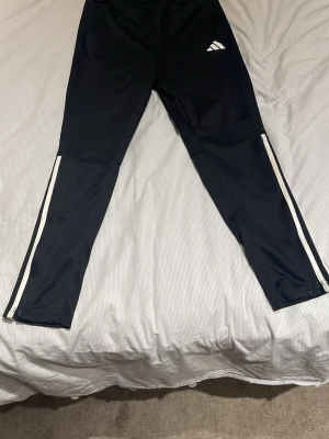 Svarta träningsbyxor från Adidas - Säljer ett par svarta träningsbyxor från Adidas med vita ränder längs benen och dragkedjor vid bensluten. Byxorna har en smal passform och är tillverkade i ett stretchigt funktionsmaterial som passar perfekt till träning eller sport.