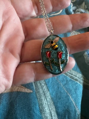 Halsband med bi och rosor - Unikt halsband med ett ovalt hänge i blått, dekorerat med en söt bi och två röda rosor med gröna blad. Hänget har en silverfärgad ram och kedja. Perfekt för dig som gillar naturinspirerade smycken och vill sticka ut med en färgglad accessoar.