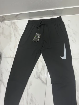 Svarta Nike mjukisbyxor med logga - Snygga svarta mjukisbyxor från Nike med stor vit Swoosh-logga på benet. Byxorna har resår i midjan med dragsko och är tillverkade i ett mjukt och lätt material. Perfekta för chill eller träning. Klassisk rak passform och diskret detalj på benet.