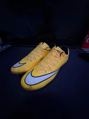 Nike Mercurial ELITE - Säljer ett par gula Nike Mercurial fotbollsskor med vit och svart swoosh på sidan. Skorna har snörning och dobbar för bra grepp på planen. Tillverkade i syntetmaterial för lätt känsla och bra bollkontroll. Perfekta för dig som vill sticka ut på fotbollsplanen.