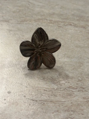 Stor silverfärgad blomring - Säljer en unik ring i silverfärgad metall med en stor blomma som motiv. Ringen har fem detaljerade blomblad och justerbar ringdel med mönstrad yta. Perfekt för dig som vill sticka ut med ett statement-smycke.