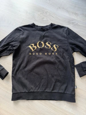 Svart sweatshirt från Hugo Boss - Svart sweatshirt med rund hals från Hugo Boss. Stor gul BOSS-logga framtill och ribbade muddar vid ärmslut och nederkant. Klassisk och stilren design i mjukt material, perfekt för en avslappnad look.
