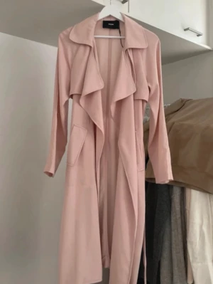 Ljusrosa trenchcoat från bikbok - Trendig ljusrosa trenchcoat från bikbok med långa ärmar, bred krage. Knytband i midjan och två stora fickor. Tillverkad i ett mjukt, tunt tyg som ger ett avslappnat intryck.