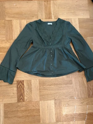 Grön blus med broderade detaljer - Mörkgrön blus från meet-me-there, v-ringning och broderade spetsdetaljer längs halsringningen och ärmsluten. Blusen har knappar framtill och vida långärmade ärmar. Sitter som en XS. Aldrig använd!