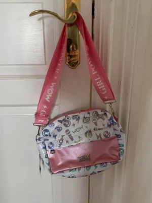 Axelväska med rosa band och tryck - Axelväska med brett rosa axelband där det står 'GIRL POWER' och 'WOW'. Väskan är vit med färgglada symboler och har en rosa metallicficka framtill. Ovalt format och dragkedja upptill. Perfekt för dig som vill sticka ut med coola detaljer.