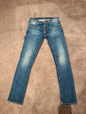 Blå raka jeans från Nudie Jeans - Säljer ett par klassiska blå jeans från Nudie Jeans med raka ben och snygga slitningar. Jeansen har fem fickor, ljusare partier på låren och bakfickor med den ikoniska Nudie-sömmen. Tillverkade i bomull med en skön passform.