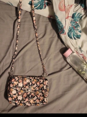 Blommig axelväska med kedjerem - Snygg axelväska med blommigt mönster i rosa, beige och grönt på svart botten. Väskan har en lång kedjerem med tygdetaljer och ett silverfärgat spänne framtill. Perfekt för dig som gillar romantisk och trendig stil.