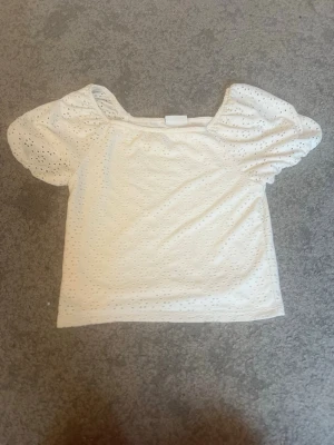 Vit blus med puffärm och broderi - Säljer en söt vit blus med puffiga korta ärmar och broderade hålmönster. Blusen har en fyrkantig halsringning och är gjord i bomull, vilket gör den både bekväm och luftig. Perfekt för dig som gillar romantiska detaljer och stilrena toppar💓💓