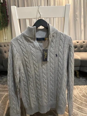 Grå kabelstickad tröja Polo Ralph Lauren - Snygg grå kabelstickad tröja från Polo Ralph Lauren med halvzip och hög krage. Tröjan har ett klassiskt broderat logotyp på bröstet och ribbstickade muddar. Perfekt för lager-på-lager och en stilren look.                                          Passar både till folk med storlek S/M