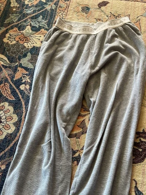 Grå loungebyxor från Lindex - Mysiga grå loungebyxor från Lindex i ribbad, mjuk ekologisk bomull. Byxorna har bred resår i midjan med texten 'SLEEP PLAY REPEAT' och avslappnad passform. Perfekta för chill dagar hemma.