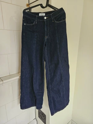 Mörkblå wide leg jeans från Lindex - Snygga mörkblå jeans från Lindex med wide leg passform och klassiska fickor både fram och bak. Jeansen är midwaist och är gjorda i denim med kontrastsömmar. Perfekta för dig som gillar en avslappnad och trendig stil.