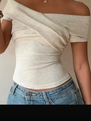 Beige offshoulder topp - Trendig beige offshoulder topp med asymmetrisk design och tight passform. Toppen är ärmlös och har en snygg drapering över bröstet. Perfekt att styla med jeans eller kjol för en cool look.