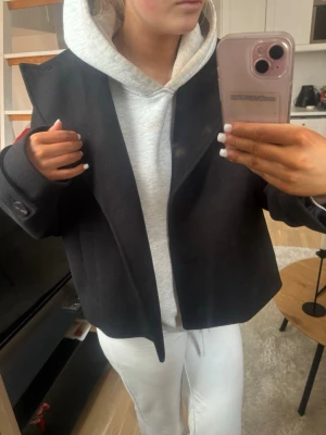 Svart skepparkavaj med knappar - Stilren svart skepparkavaj med bred krage och stora knappar vid ärmsluten. Jackan har en boxig, lite croppad passform och är perfekt att slänga över en hoodie för en chill look. Tillverkad i ett tjockare material som känns både klassiskt och trendigt.