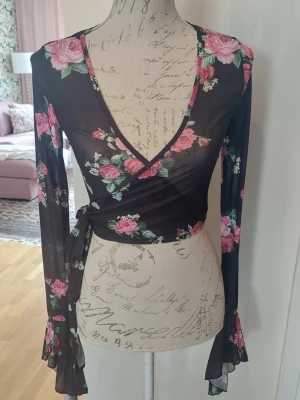 Svart blommig omlottopp med knyt - Snygg svart omlottopp med rosa blommor och långa ärmar. Toppen är transparent, har djup v-ringning och knyt i sidan. Ärmarna har volangdetaljer vid ärmsluten. 