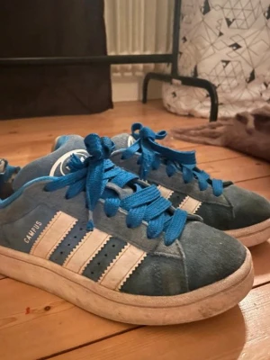 Blå Adidas Campus sneakers - Säljer ett par blå Adidas Campus sneakers med klassiska vita ränder på sidorna och blå skosnören. Skorna har en låg profil och är gjorda i mocka med vit sula. Perfekta för dig som gillar retrostil och streetwear.