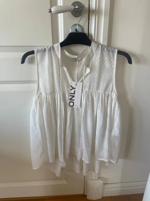 Vit ärmlös blus från ONLY - Superfin vit blus från ONLY med diskret prickigt mönster och volangdetaljer. Blusen är ärmlös och har en luftig, flowy passform med v-ringning och liten krage. Perfekt för en fräsch och somrig look.