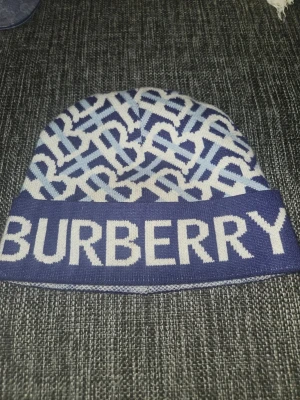 Blåvit Burberry mössa med logga - Snygg mössa från Burberry i blått och vitt med stor logga framtill och mönster över hela mössan. Tillverkad i mjukt stickat material som håller dig varm och ger en trendig look. Perfekt accessoar för att lyfta din outfit.