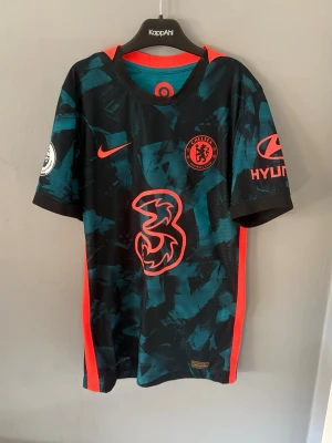 Chelsea FC bortatröja Nike - Chelsea FC bortatröja med Chilwell 21 på ryggen. Tröjan är svart med turkosa mönster och röda detaljer, samt stora vita tryck. Nike-logga, klubbmärke och sponsorer på bröst och ärmar. Perfekt för fotbollsfans som vill sticka ut på läktaren.