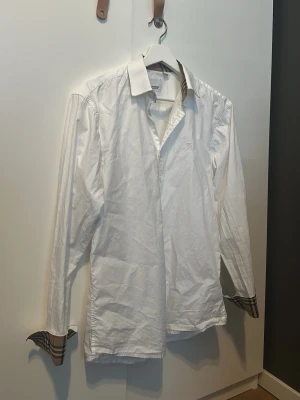 Burberry skjorta - Klassisk vit skjorta från Burberry med diskret broderad logga på bröstet och ikoniska rutiga detaljer på insidan av kragen och manschetterna. Skjortan har lång ärm och knappar framtill. Perfekt för dig som gillar stilrena plagg med exklusiv känsla. Inga hål eller defekter. 