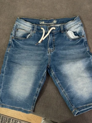 Blå jeansshorts med snörning LAB - Säljer ett par blå jeansshorts från LAB med vit snörning i midjan och klassiska fem fickor. Shortsen har en tvättad look och är tillverkade i bomullsmaterial. Perfekta för sommaren och har en avslappnad passform.