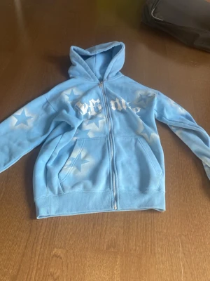 Blå zip hoodie med stjärnor FSBN - Ljusblå hoodie från FSBN i storlek XS med dragkedja framtill och vita stjärnor samt text i graffiti-stil över bröstet. Hoodien har kängurufickor och ribbade muddar. Perfekt för en avslappnad streetwear-look.