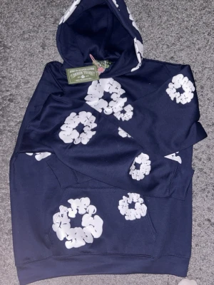 Mörkblå hoodie från Denim Tears - Snygg mörkblå hoodie från Denim Tears med vita blommönster över hela tröjan. Hoodien har en klassisk huva och magficka framtill. Perfekt för dig som vill sticka ut med en unik och trendig stil.