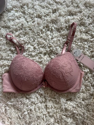Rosa push-up bh med spetsdetaljer - Söt rosa push-up bh med vadderade kupor och vackra spetsbroderier i blommönster. Mellan kuporna sitter en liten rosett med pärldetalj. Justerbara axelband och hakknäppning bak. Perfekt för dig som vill ha både komfort och en feminin look.