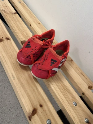 Röda Adidas fotbollsskor - Säljer ett par röda fotbollsskor från Adidas med klassiska vita ränder på sidan och svarta detaljer på tungan. Skorna har snörning och en låg profil, perfekta för snabba rörelser på planen. Materialet är syntet med textilfoder för skön känsla.