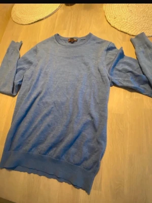 Blå stickad tröja i merinoull - Snygg blå stickad tröja från J. Lindeberg i mjuk merinoull. Tröjan har rund halsringning, långa ärmar och ribbade muddar vid ärmslut och nederkant. Perfekt för lager på lager och enkel att matcha med olika outfits.