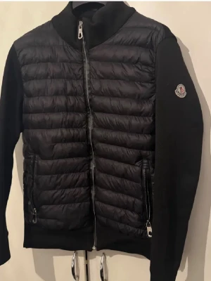 Svart dunväst från Moncler - Snygg svart dunväst från Moncler med quiltad front och två dragkedjeförsedda fickor. Västen har ribbstickad krage och Moncler-logga på ärmen. Materialet är en mix av dun och textil, vilket ger en sportig och modern look.