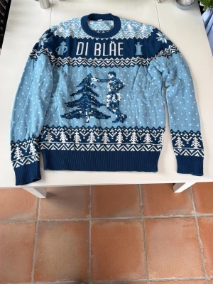 Blå stickad tröja DI BLÅE - Säljer en blå stickad tröja med texten 'DI BLÅE' och motiv av en gran och en person. Tröjan har vita och mörkblå detaljer, snöliknande prickar och mönster längs ärmar och nederkant. Perfekt för dig som gillar unika stickade plagg.