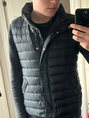 Svart dunväst med hög krage - Snygg svart dunväst med hög krage och tryckknappar framtill. Västen har en quiltad design med horisontella sömmar och två sidofickor. Enkel och stilren look som passar till det mesta.