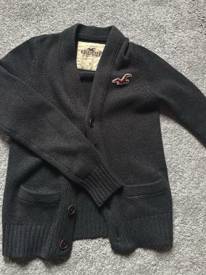 Svart kofta från Hollister - Mysig mörkgrå kofta från Hollister med två framfickor och bruna knappar. Klassisk v-ringad design och broderad logga i vinrött och vitt på bröstet. Perfekt att slänga över en t-shirt för en chill look.