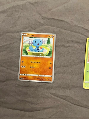 Pokémonkort Wooper japansk - Japanskt Pokémonkort med Wooper, orange bakgrund och färgglad illustration av Wooper i gräs. Kortet har japansk text och visar Wooper med blå kropp och lila detaljer. Perfekt för samlare eller Pokémonfans.