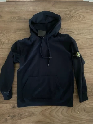 Mörkblå hoodie från Stone Island - Snygg mörkblå hoodie från Stone Island med klassisk logga på ärmen. Hoodien har huva med dragsko och en stor magficka framtill. Perfekt för dig som gillar streetwear och vill ha en stilren look. Materialet känns mjukt och skönt mot huden.