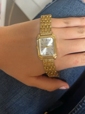 Guldfärgad fyrkantig klocka  - Stilren guldfärgad klocka med fyrkantig urtavla och länkarmband i metall. Urtavlan är silverfärgad med romerska siffror och minimalistiska markeringar. Perfekt accessoar för dig som gillar klassisk och tidlös design. Kommer inte ihåg vart den är köpt tror ur&penn