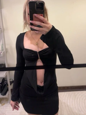 Svart långärmad cut-out klänning - Trendig svart kortklänning från Glamine med lång ärm och djup cut-out framtill. Klänningen har en snygg detalj med ring i brösthöjd och tight passform som framhäver figuren. Perfekt för dig som vill sticka ut med en unik och modern look.