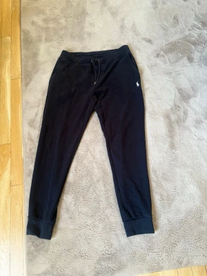 Svarta mjukisbyxor från Polo Ralph Lauren - Svarta mjukisbyxor från Polo Ralph Lauren med dragsko i midjan och ribbade muddar vid bensluten. Byxorna har en liten vit logga broderad på vänster sida och är tillverkade i mjukt bomullsmaterial. Perfekta för en avslappnad och stilren look.
