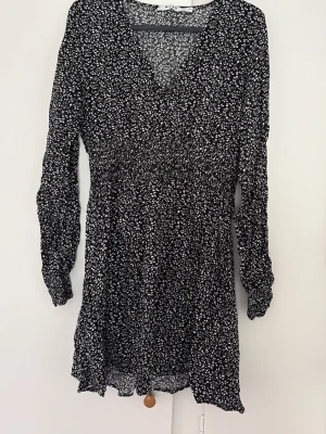 Svartvit blommig klänning NA-KD - Supersöt svartvit blommig kortklänning från NA-KD med v-ringning och långa ärmar. Klänningen har smock i midjan för en snygg passform och är gjord i ett lätt och luftigt material. Perfekt för dig som gillar en feminin och trendig stil.