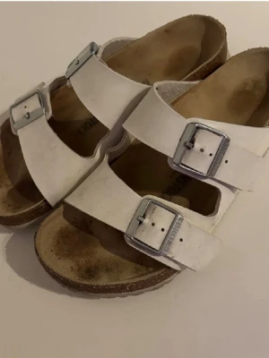 Vita Birkenstock  - Klassiska vita sandaler från Birkenstock med två breda remmar och stora silvriga metallspännen. Fotbädden är formad för extra komfort och sulan är i kork. Perfekta för en avslappnad och trendig stil.