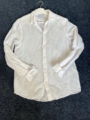 Vit linneskjorta från Pier One - Klassisk vit skjorta från Pier One i svalt linnematerial. Skjortan har lång ärm, knappar framtill och en avslappnad passform. Perfekt för en stilren och enkel look. Snygg att bära både uppknäppt och stängd.