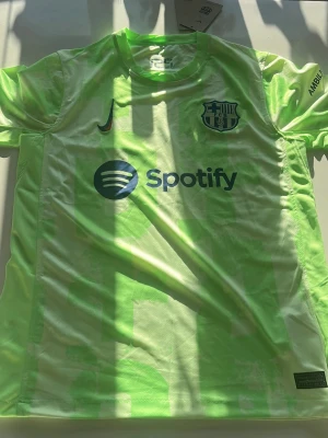 Neongul FC Barcelona matchtröja Nike - Säljer en neongul FC Barcelona matchtröja från Nike med klubbmärke på bröstet och Spotify-logga på framsidan. Tröjan har UNHCR-logga på ryggen och är tillverkad i lätt, ventilerande syntetmaterial. Perfekt för dig som vill sticka ut på planen.
