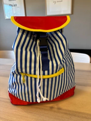 Randig ryggsäck med färgglada detaljer - Cool ryggsäck med blå och vita ränder, röd botten och lock samt gula detaljer. Praktisk dragkedja framtill och snörning upptill. Justerbara blå axelband. Perfekt för dig som gillar färg och mönster!