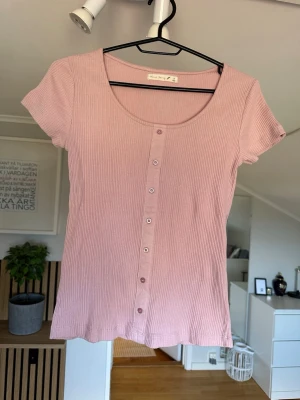 Ljusrosa ribbad topp med knappar - Supersöt ljusrosa ribbad topp från Black Story med korta ärmar och knappar framtill. Toppen har en rundad urringning och är tillverkad i mjuk bomullsmix som känns skön mot huden. Perfekt att styla med jeans eller kjol för en chill look.